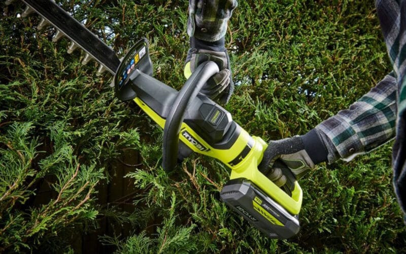 Två händer som håller en Ryobi häcksax RY36HTX65A-0 mot en grön häck