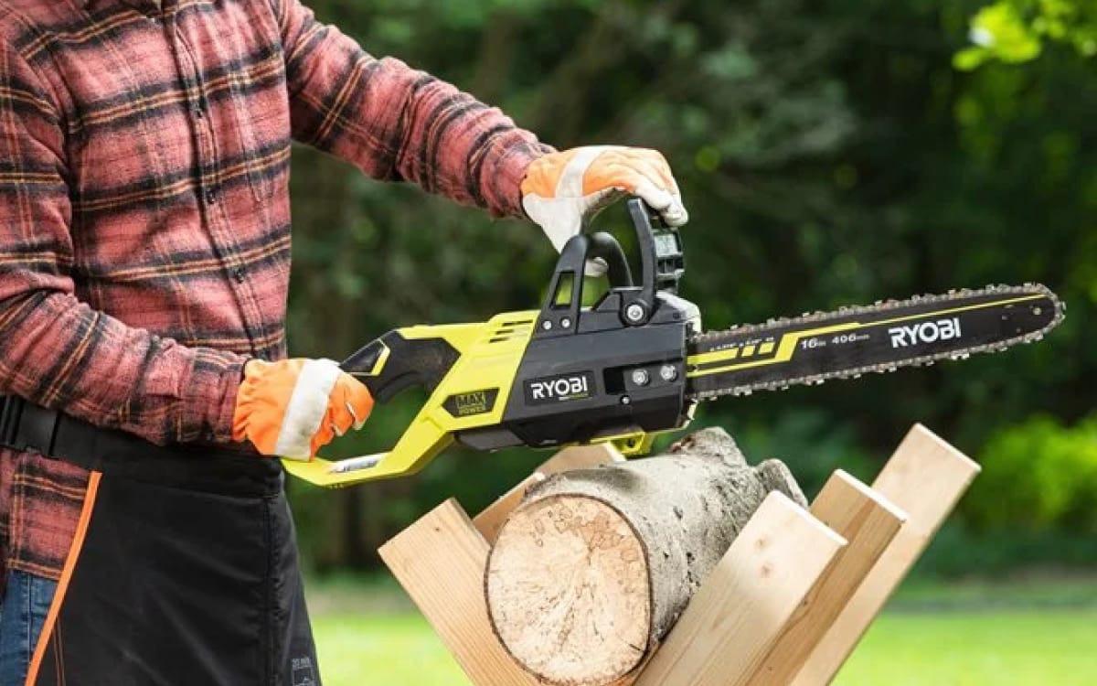 En person håller i en RYOBI kedjesåg RY36CSX40B-0 på en stock med grön gräsmatta och träd i bakgrunden